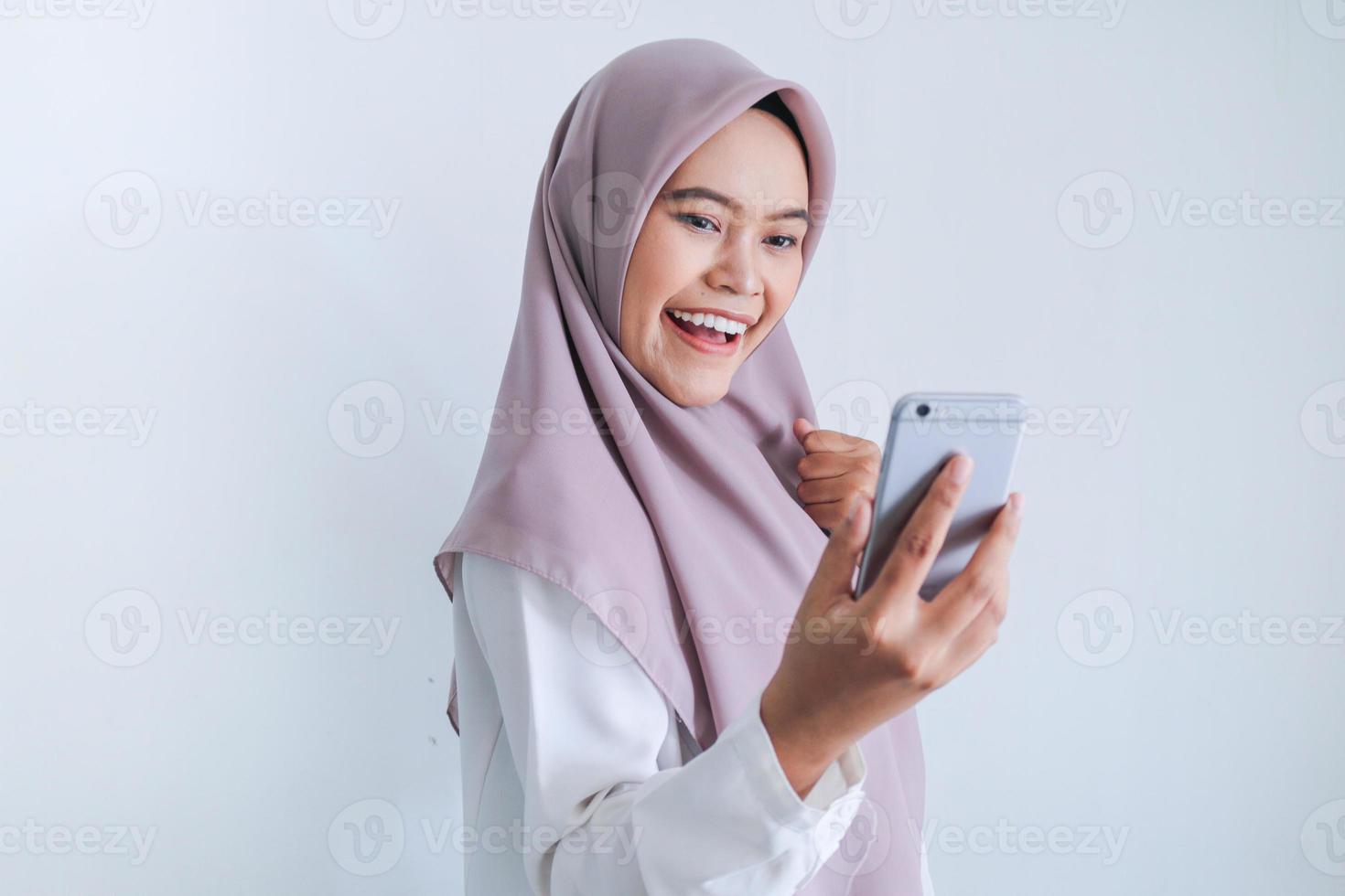 jonge aziatische islamvrouw die een hoofddoek draagt, is blij en opgewonden om te vieren wat ze op de smartphone ziet. Indonesische vrouw op grijze achtergrond foto