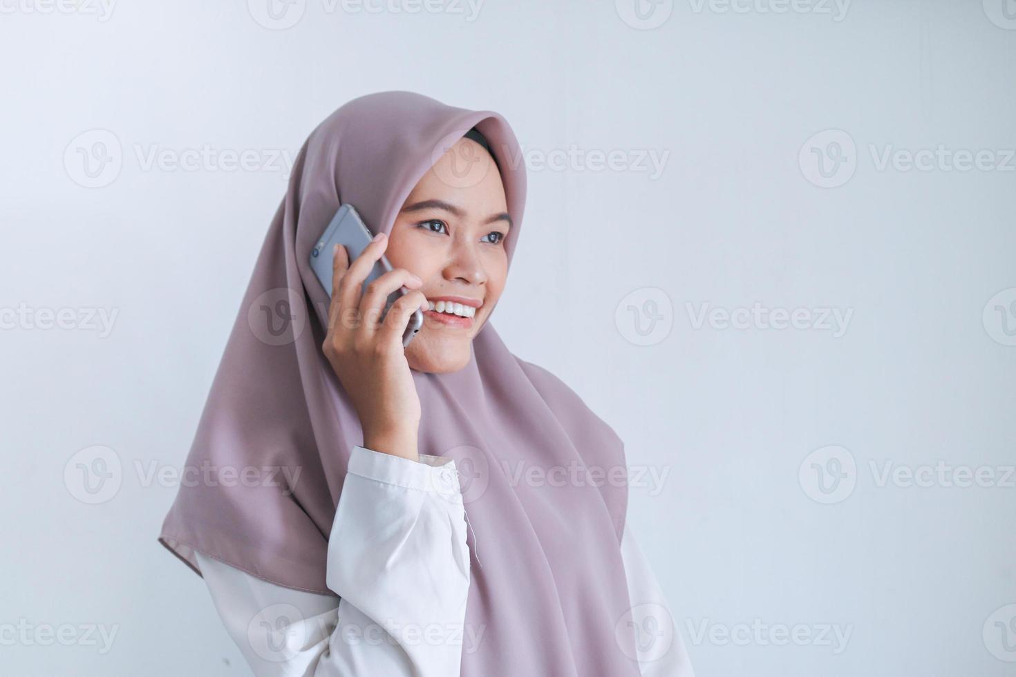 jonge aziatische islamvrouw die een hoofddoek draagt, is blij en glimlacht wanneer ze aan de telefoon praat. Indonesische vrouw op grijze achtergrond foto