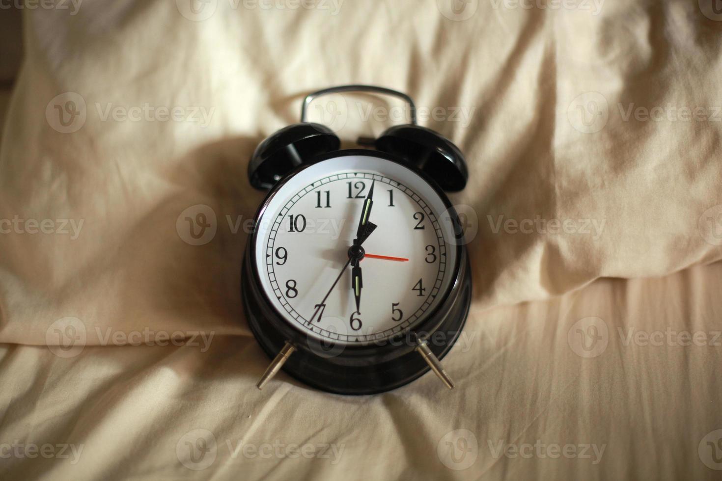 bovenaanzicht van klassieke zwarte wekker met zes uur met een bed op de achtergrond, wakker worden in de ochtend concept foto