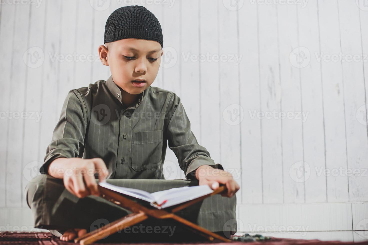 aziatische moslimjongen die traditioneel kostuum genaamd gamis draagt en het heilige boek al-koran op de gebedsmat leest foto