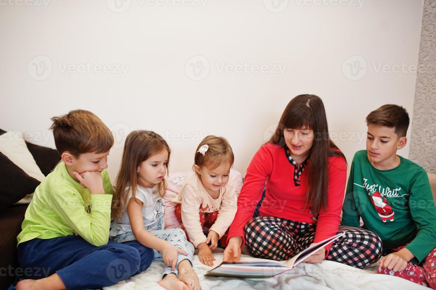 gelukkige grote familie heeft samen plezier in de slaapkamer. grote familie ochtend concept. moeder met vier kinderen draagt thuis een pyjama leesboek in bed. foto