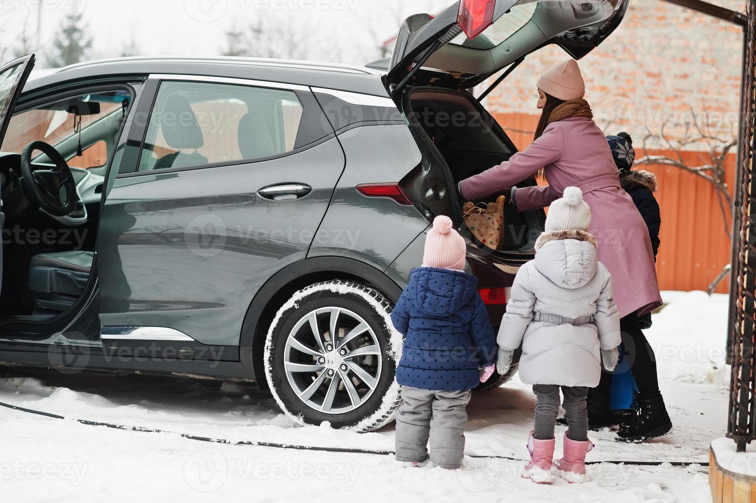 jonge vrouw met kinderen houdt ecotassen vast en laadt elektrische auto op met open kofferbak in de tuin van haar huis. foto