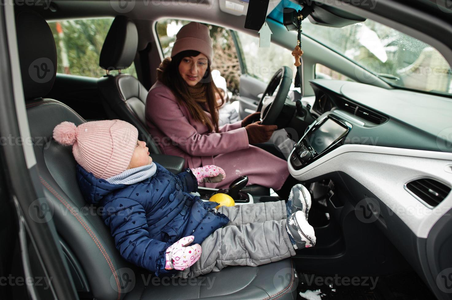 jonge moeder met dochtertje van de baby zit in de elektrische auto in de tuin van haar huis in de winter. foto