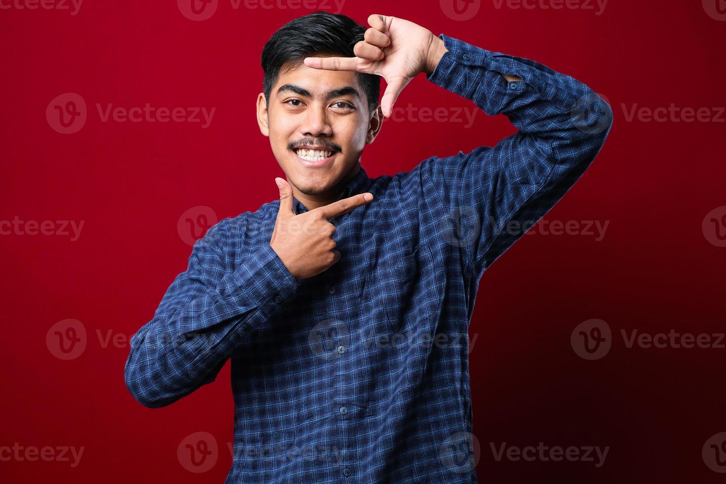 jonge knappe aziatische jongen met een casual shirt over een geïsoleerde rode achtergrond die lacht en een frame maakt met handen en vingers met een blij gezicht. foto