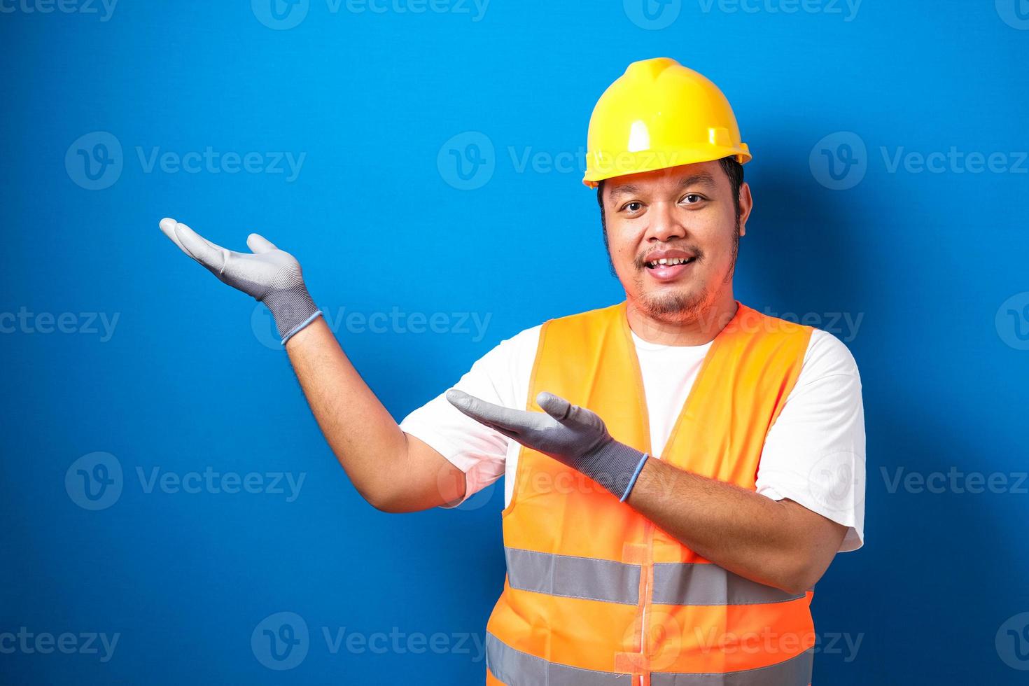dikke Aziatische arbeider die een helm draagt, presenteert iets in zijn hand terwijl hij zijwaarts kijkt foto