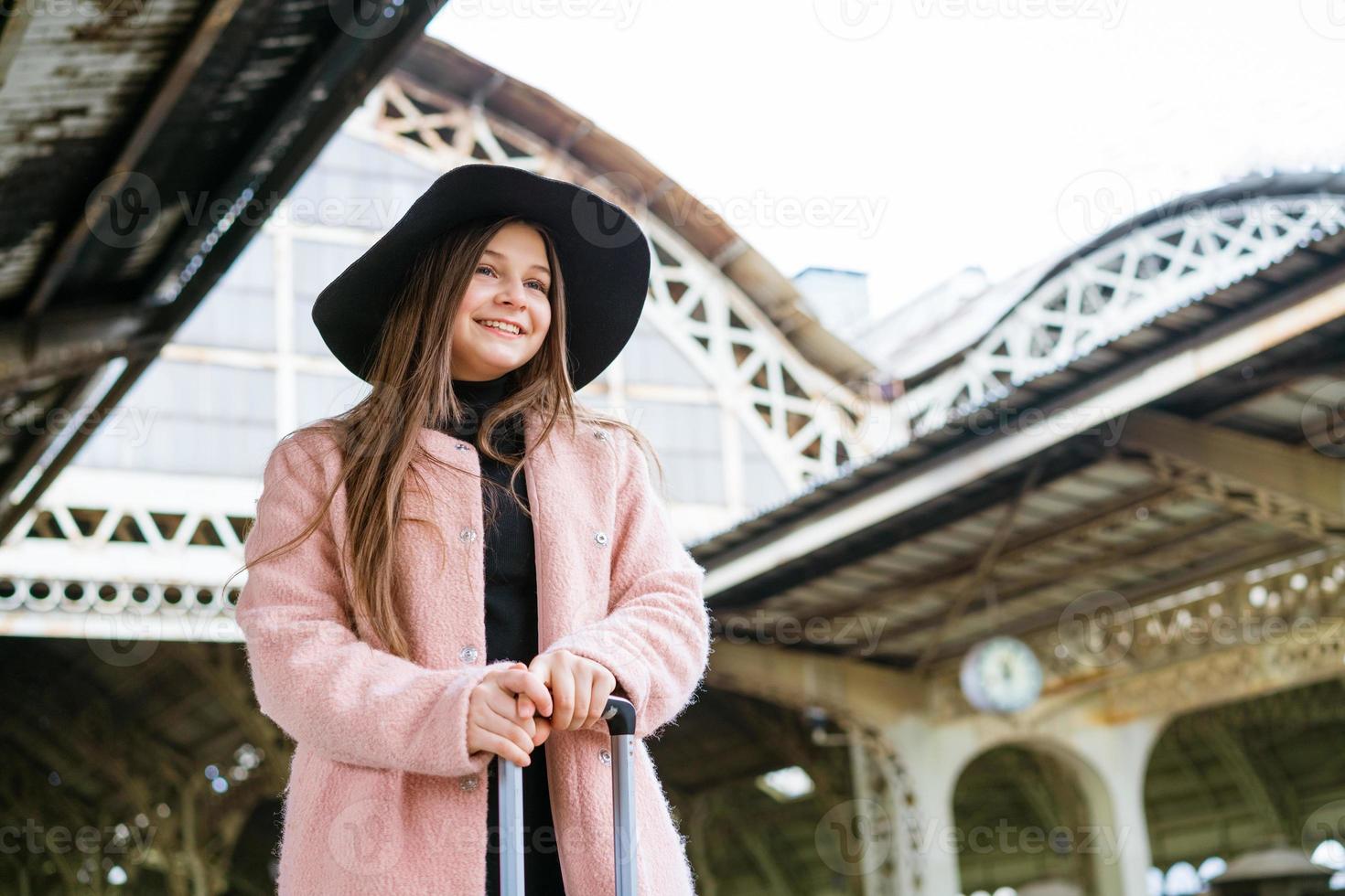 mooie jonge casual toeristische vrouw met een koffer in een roze jas te wachten foto