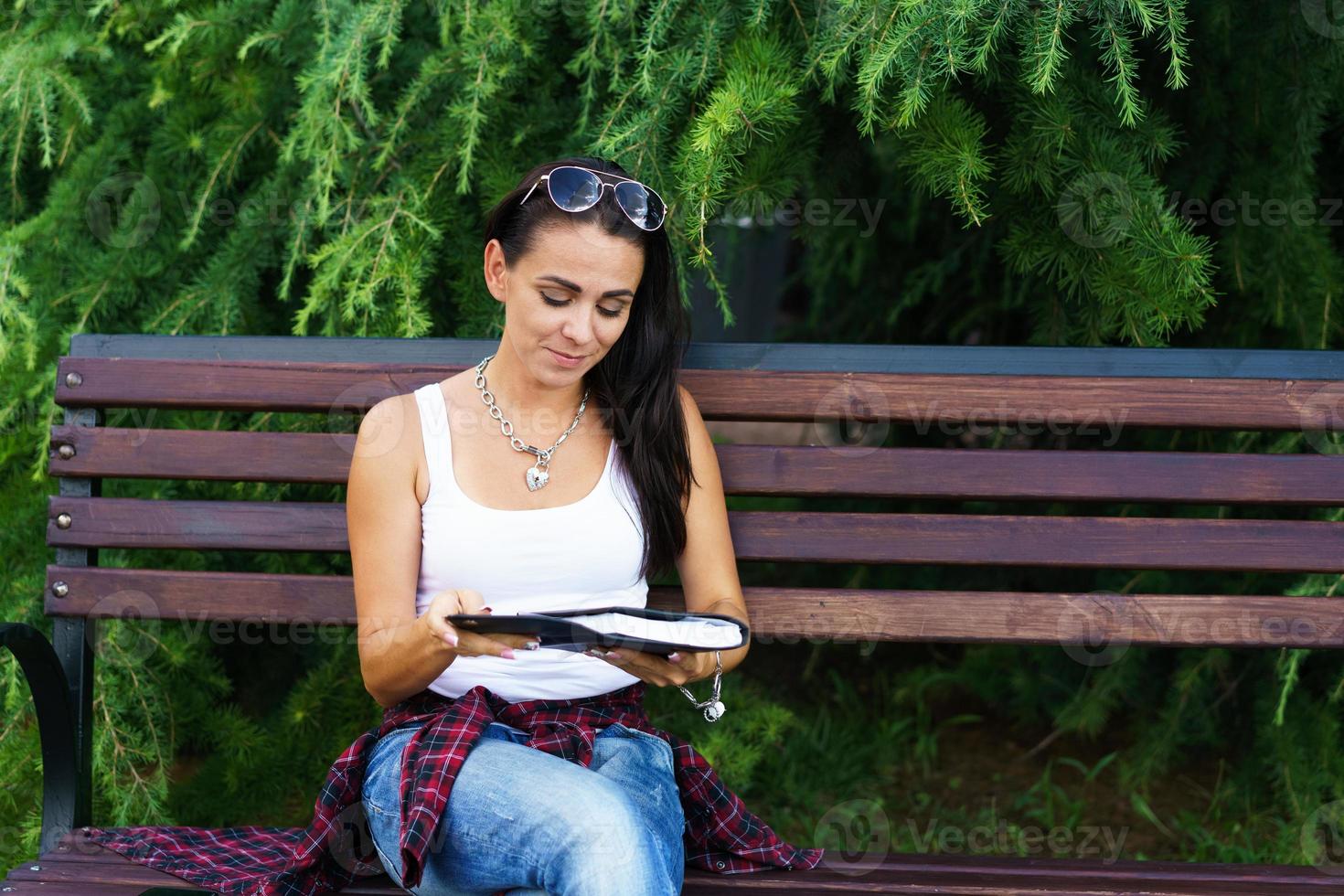 vrolijke brunette vrouw in vrijetijdskleding en zonnebril zit op een houten bankje foto