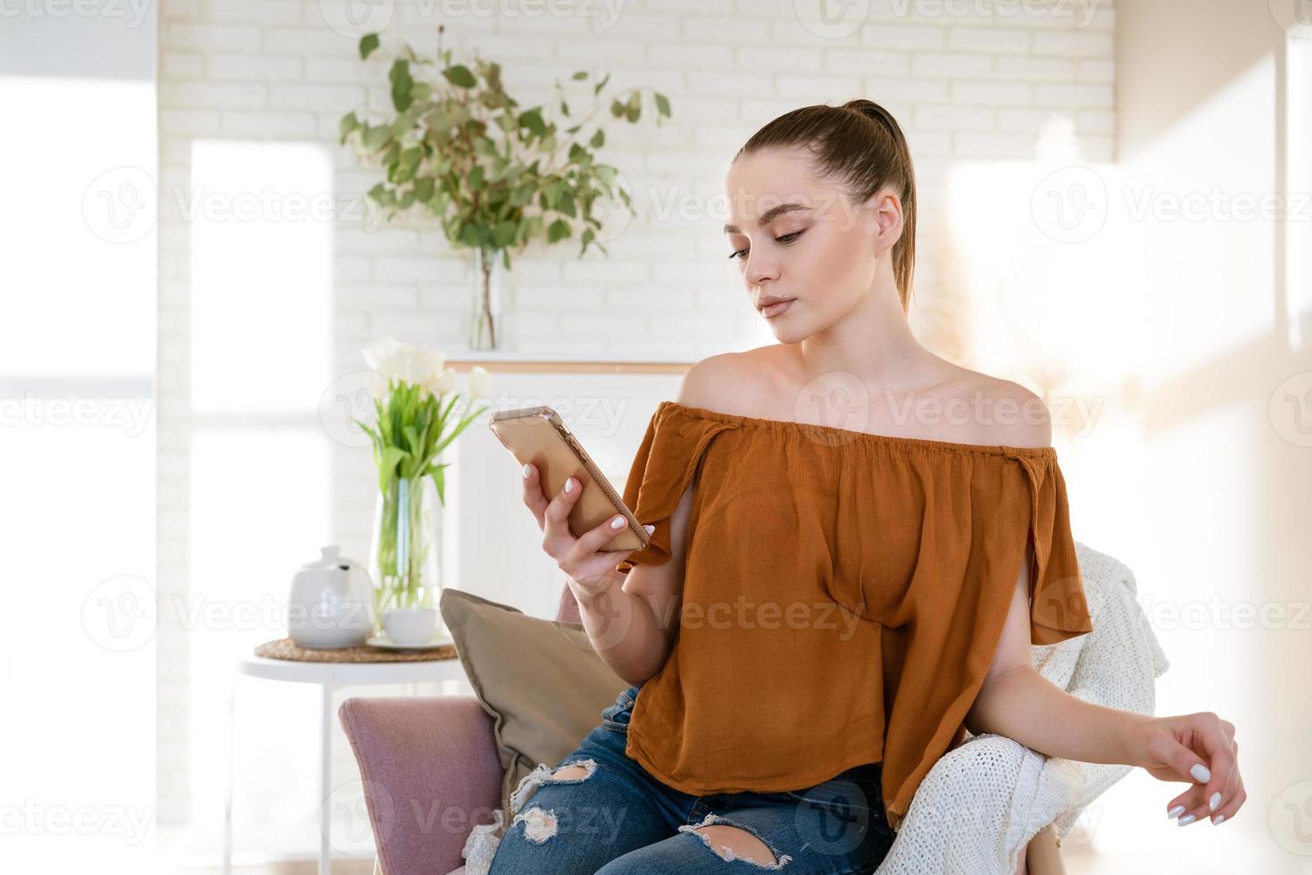 een schattig meisje zit in een licht interieur in een fauteuil met een telefoon in haar hand foto