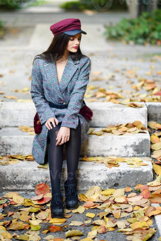 jong mooi meisje met winterjas en pet zittend op trappen vol herfstbladeren foto