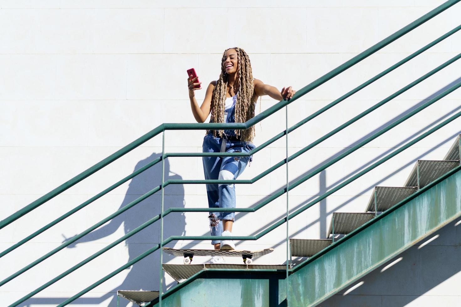 zwarte vrouw met gekleurde vlechten, kijkend naar haar smartphone met haar voeten op een skateboard. foto