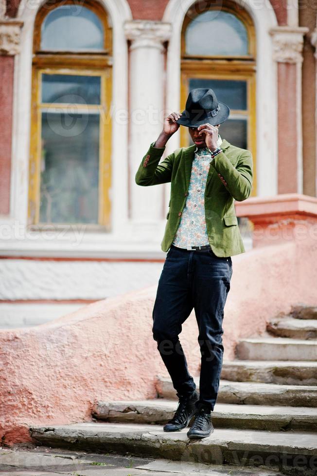 mode portret van zwarte Afro-Amerikaanse man op groen fluwelen jasje en zwarte hoed verblijf op trappen achtergrond oud herenhuis. verticale foto