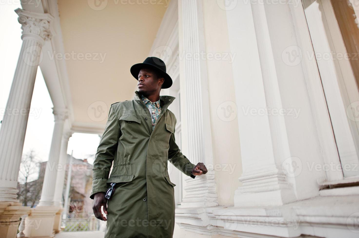 mode portret van zwarte Afro-Amerikaanse man op groene jas mantel en zwarte hoed, achtergrond geel herenhuis foto