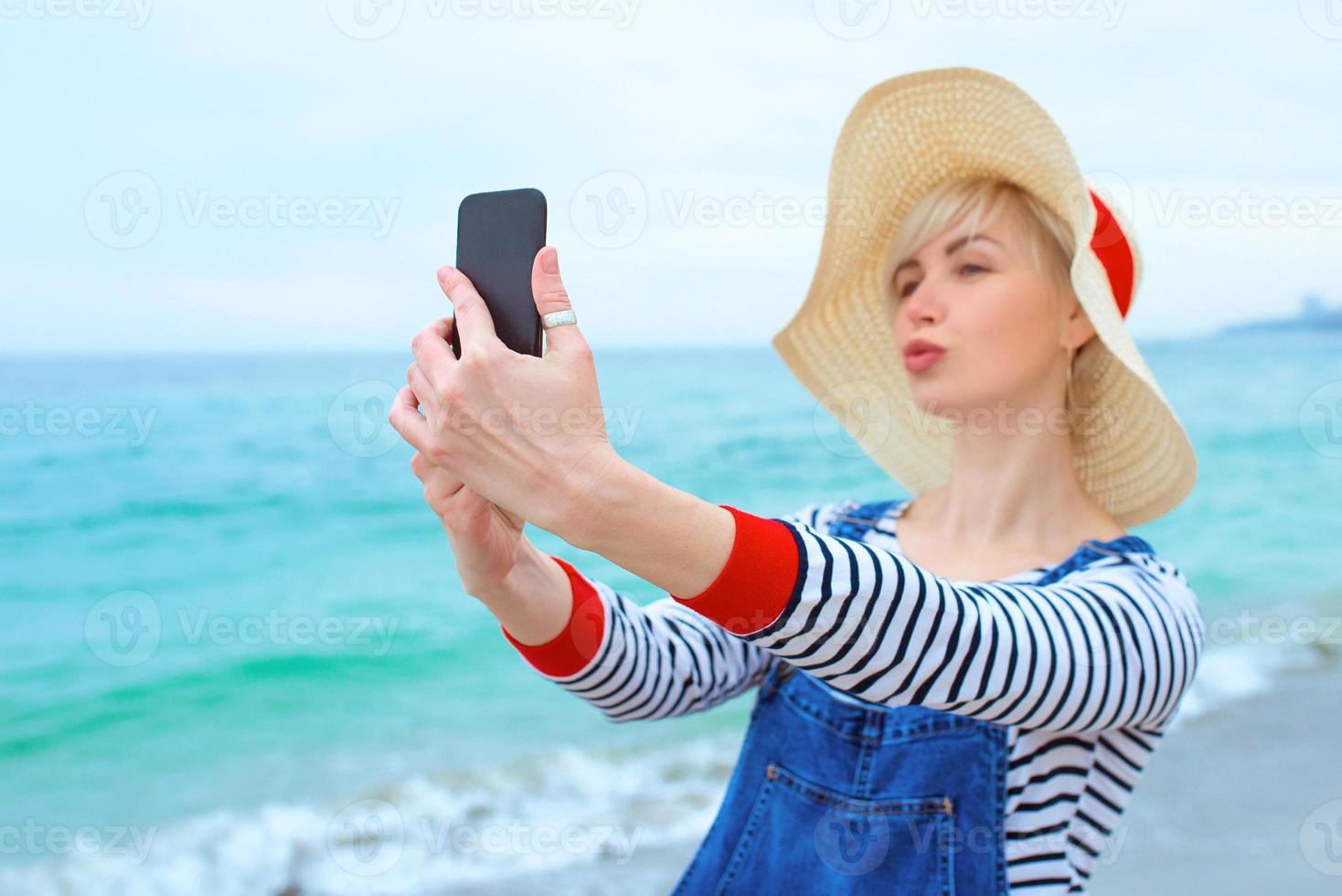 mooie jonge blonde blanke vrouw op vakantie in strohoed, gestreepte blouse en denim overall die selfie op smartphone maken door de verbazingwekkende blauwe zee-achtergrond foto