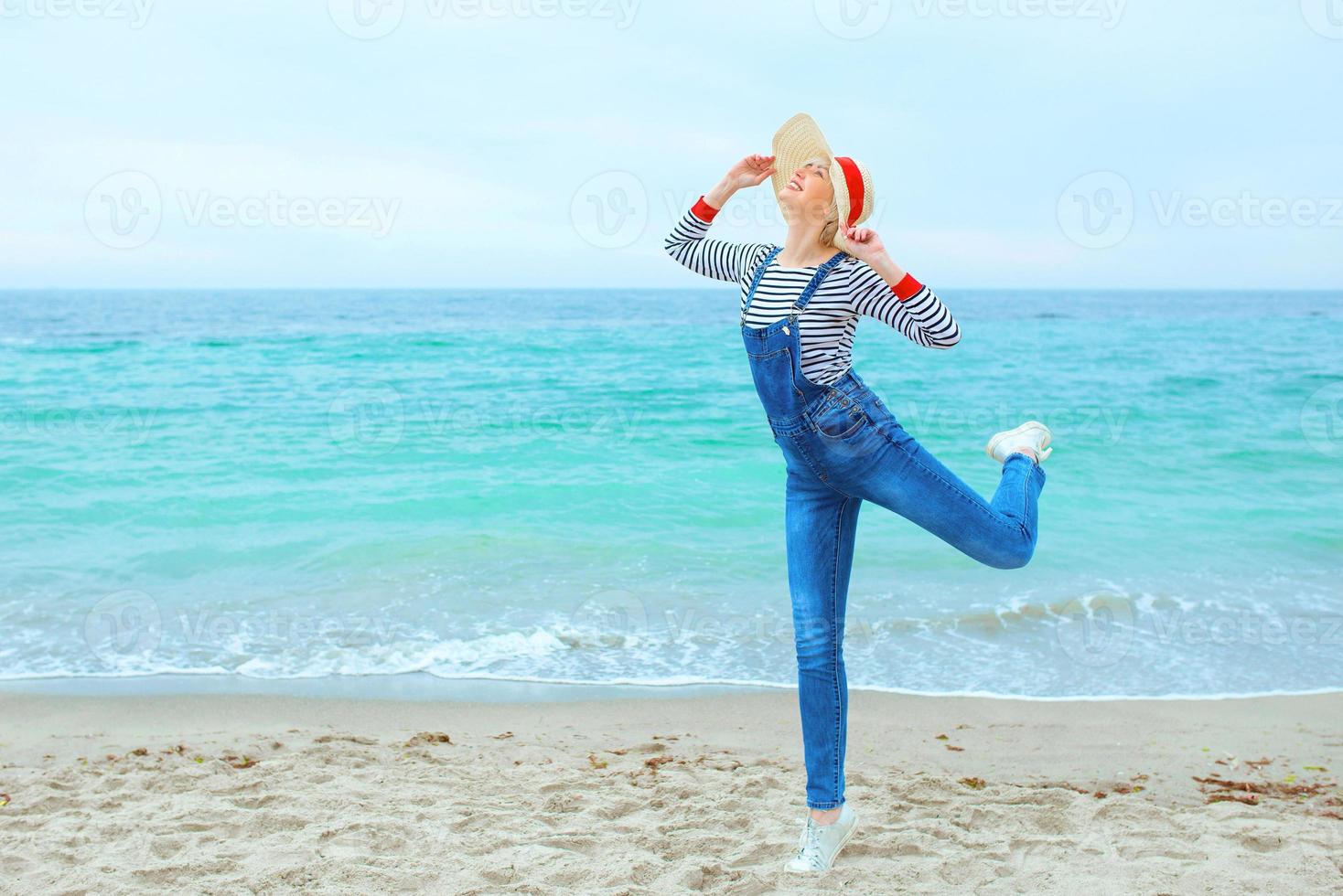 mooie jonge blonde blanke vrouw op vakantie in gestreepte blouse, sneakers en denim overall springen op het strand door de verbazingwekkende blauwe zee-achtergrond foto