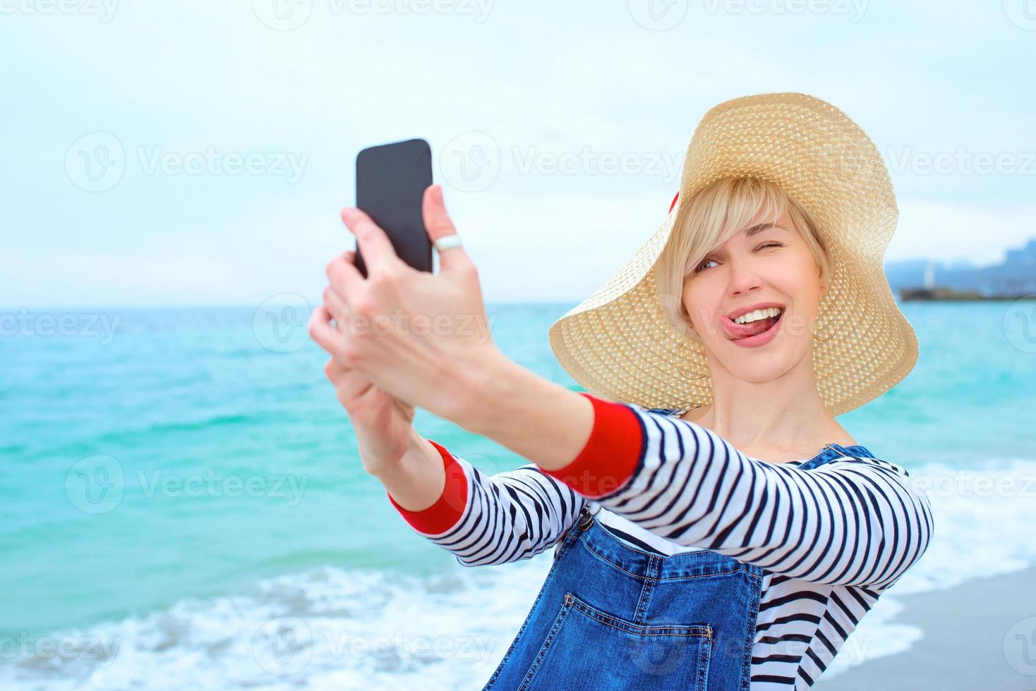 mooie jonge blonde blanke vrouw op vakantie in strohoed, gestreepte blouse en denim overall die selfie op smartphone maken door de verbazingwekkende blauwe zee-achtergrond foto