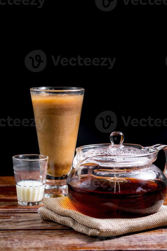 transparante theepot met hete thee op bruine doek met glas melk op houten tafel op zwarte achtergrond met kopieerruimte. beroemde drank van aziatische. gezond drankconcept foto