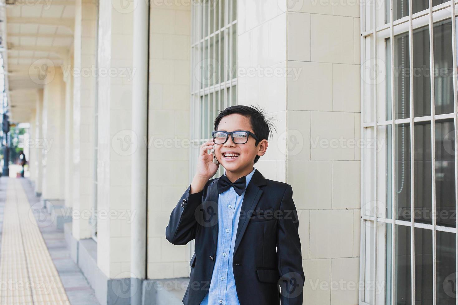 schattige aziatische jongen met een vintage zwart pak en een bril praten aan de telefoon terwijl hij lacht foto