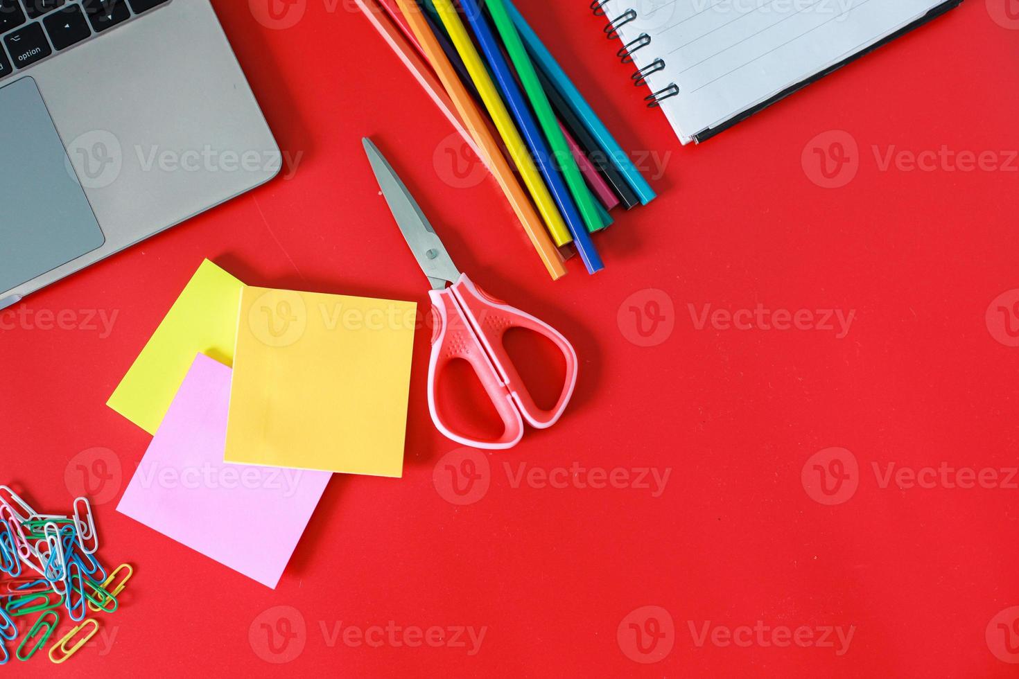 bovenaanzicht van kantoor- of schoolbenodigdheden geïsoleerd op rode achtergrond. open boek, kleurpotlood, laptop, schaar, kleurrijke paperclip en plakbriefje met vrije of kopieerruimte foto