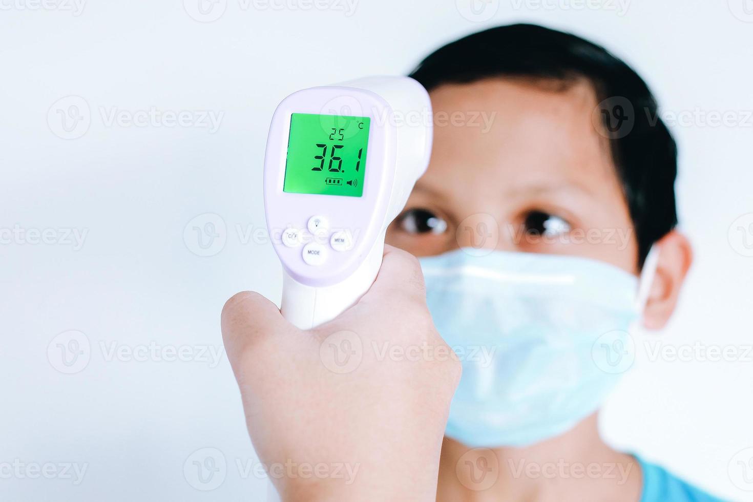 infraroodthermometer in een hand die de temperatuur meet van de Aziatische kleine jongen met beschermend chirurgisch masker op het gezicht foto