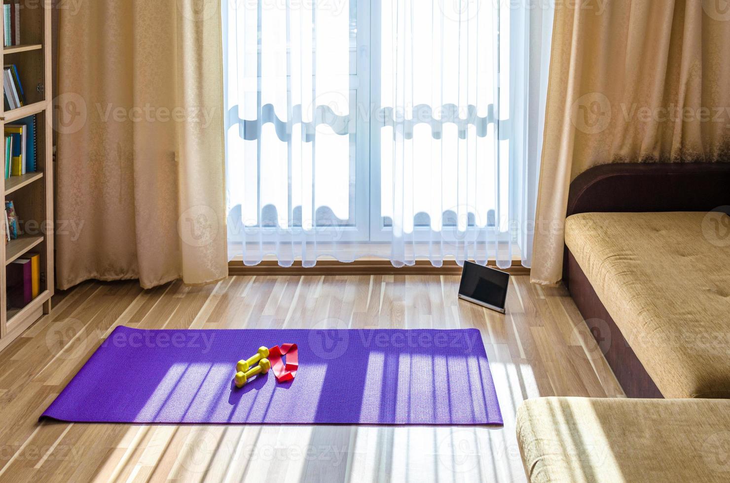 woonkamer met sportuitrusting en tablet op de vloer, boekenkast en bank bij openslaande deuren, gele halters en rode rubberen weerstandsband foto