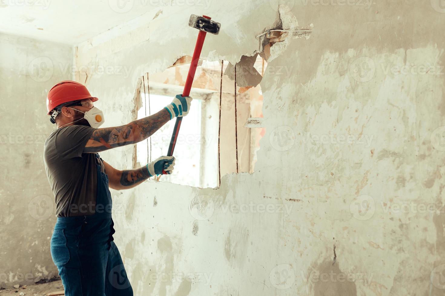 bouwer met een hamer in zijn handen breekt de betonnen muur de bouwer is gekleed in een beschermend pak en helm foto