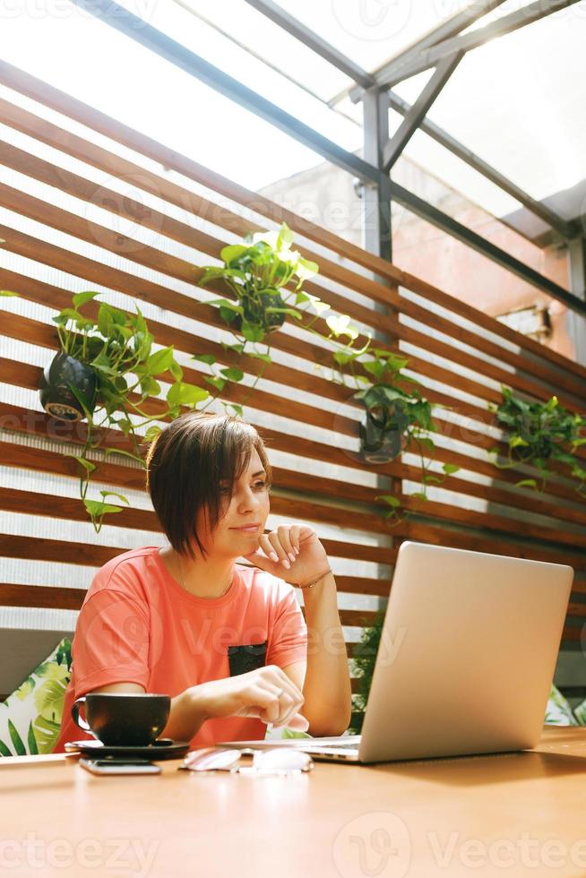 portret van zelfverzekerde volwassen professionele vrouw met een bril, een koraal t-shirt zittend op een zomerterras in café, laptopcomputer gebruikt voor werk, vrolijk lachend binnenshuis foto
