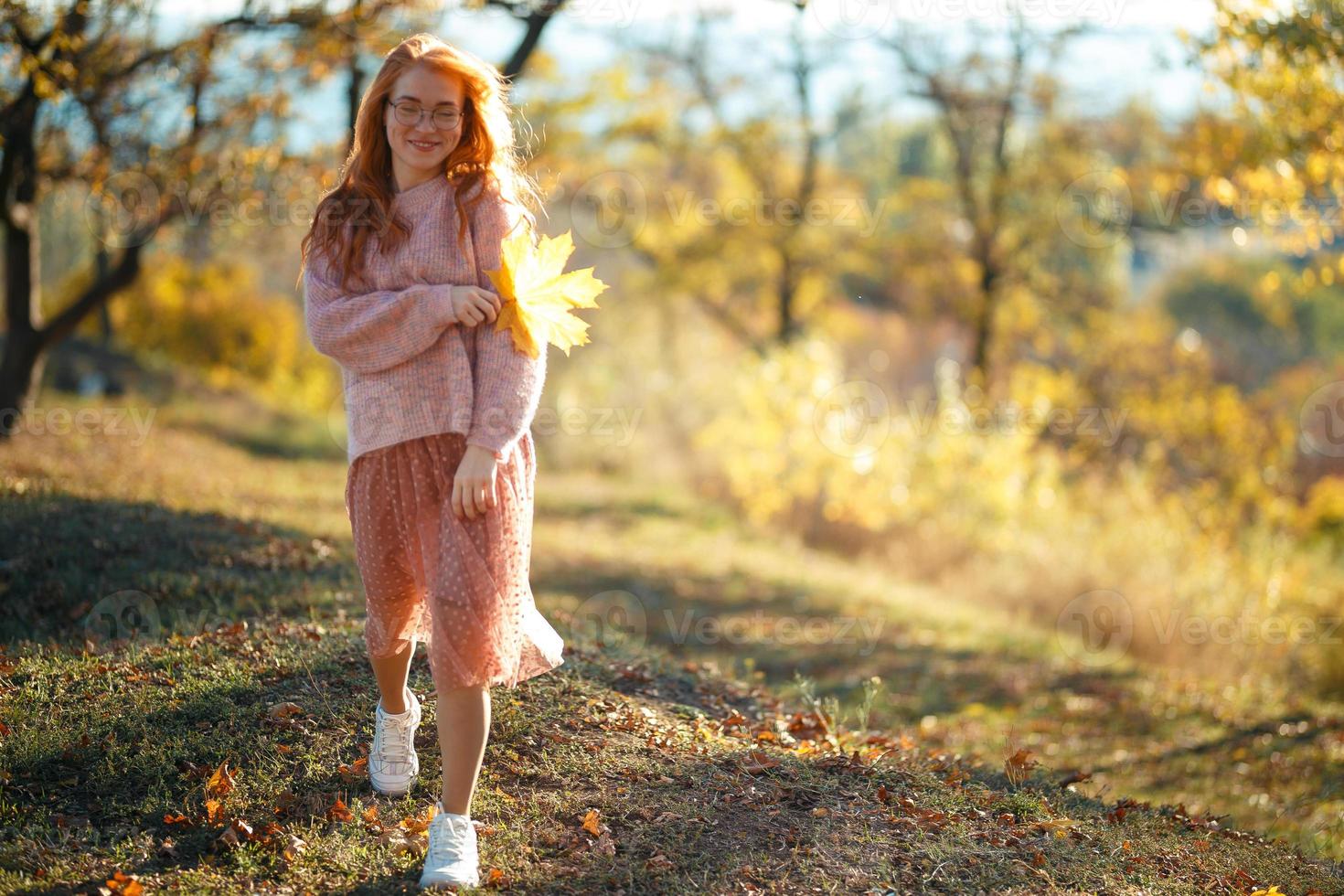 portretten van een charmant roodharig meisje met een schattig gezicht. meisje poseren in herfst park in een trui en een koraalkleurige rok. in de handen van een meisje een geel blad foto