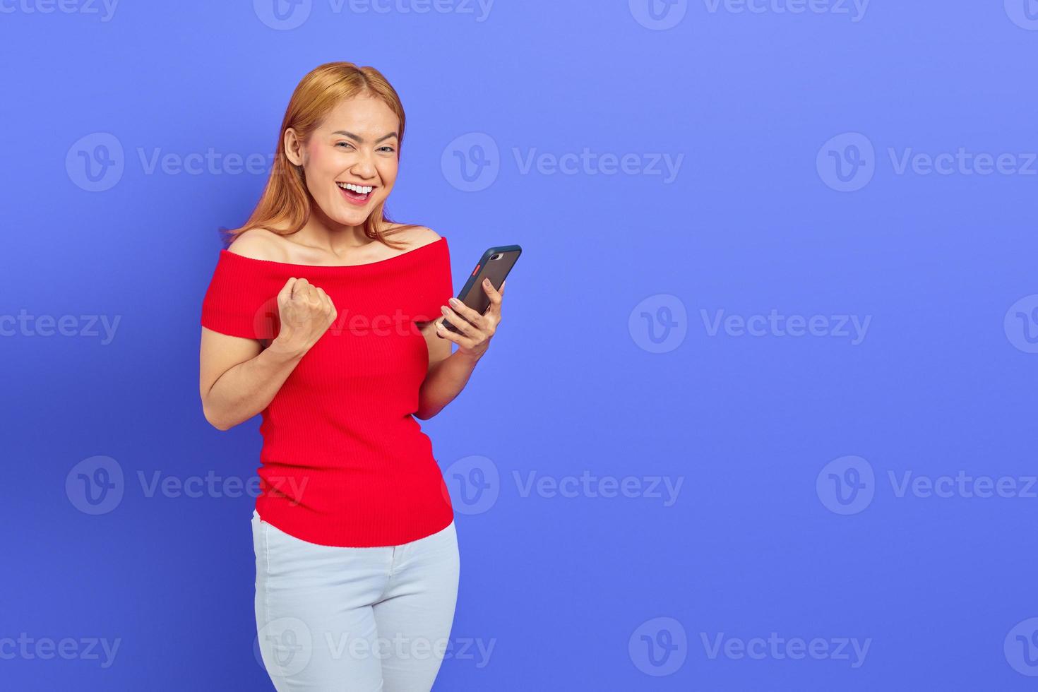 Portret van een vrolijke jonge Aziatische vrouw die een smartphone vasthoudt terwijl ze een winnend gebaar toont dat op een paarse achtergrond wordt geïsoleerd foto