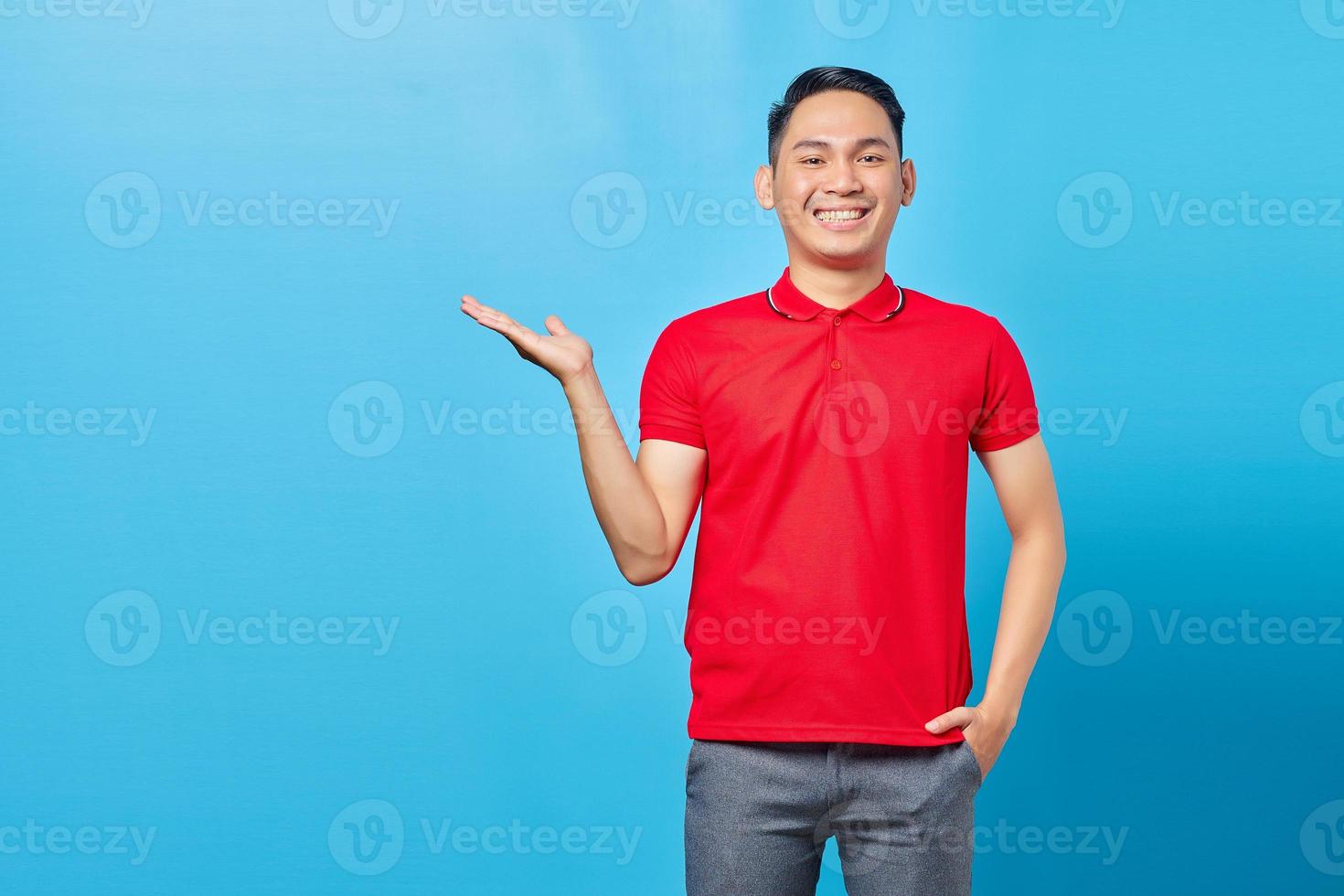 portret van een vrolijke aziatische jongeman in een rood shirt die wijst naar de ruimte met handpalmen geïsoleerd op een blauwe achtergrond foto