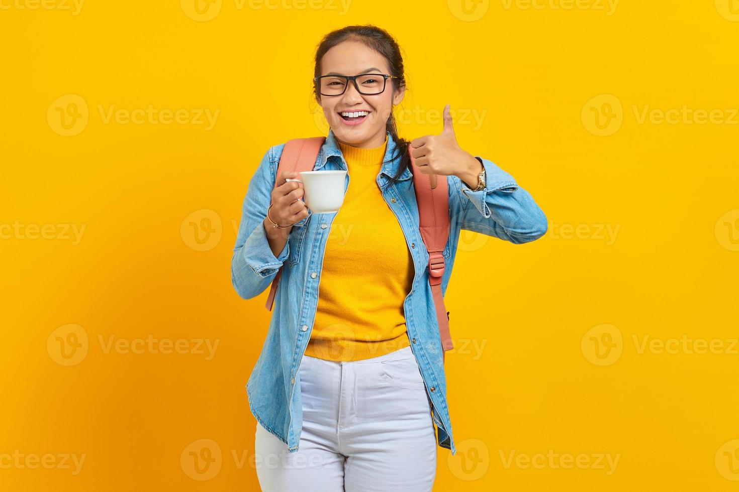 Vrolijke mooie jonge Aziatische vrouw in denim kleding met rugzak met kopje koffie en duim opdagen geïsoleerd op gele achtergrond foto