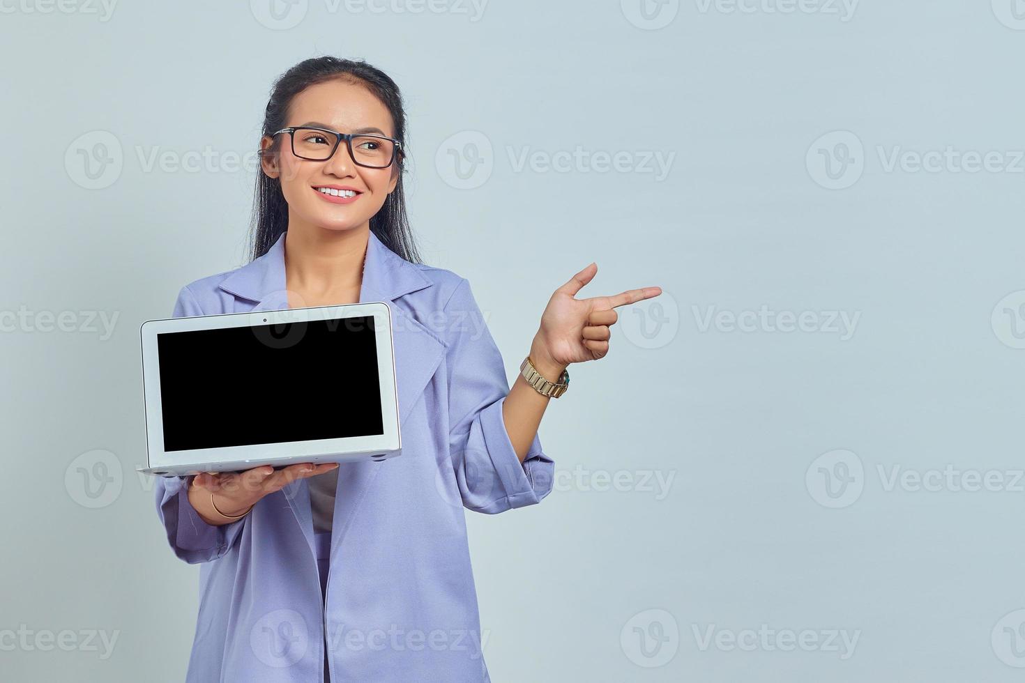 portret van een glimlachende jonge aziatische vrouw die een leeg laptopscherm toont en wijst om de ruimte te kopiëren met een vinger die op een witte achtergrond wordt geïsoleerd foto