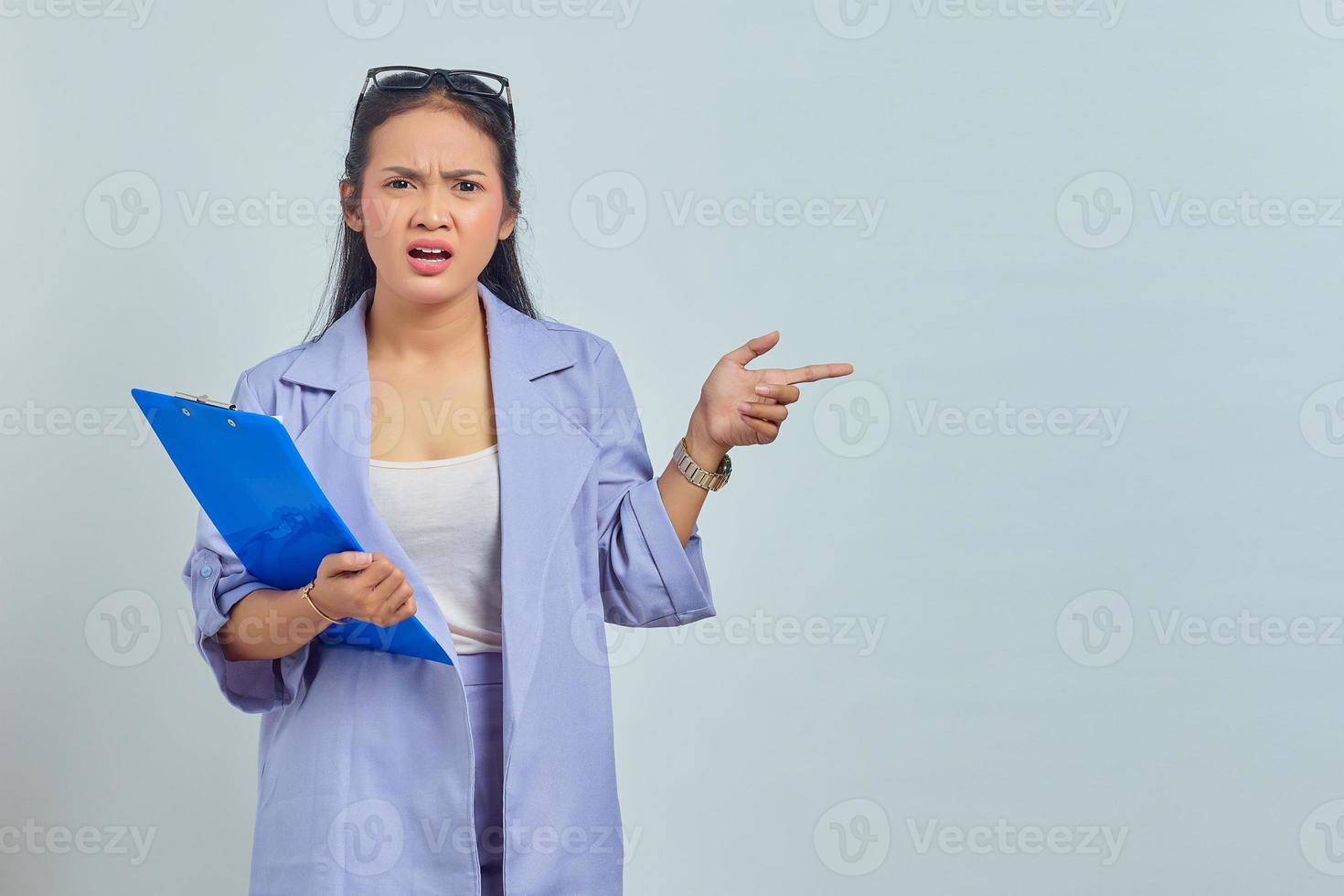 portret van geschokte jonge aziatische vrouw in pak met documentmap en wijzende vinger naar kopieerruimte geïsoleerd op paarse achtergrond foto