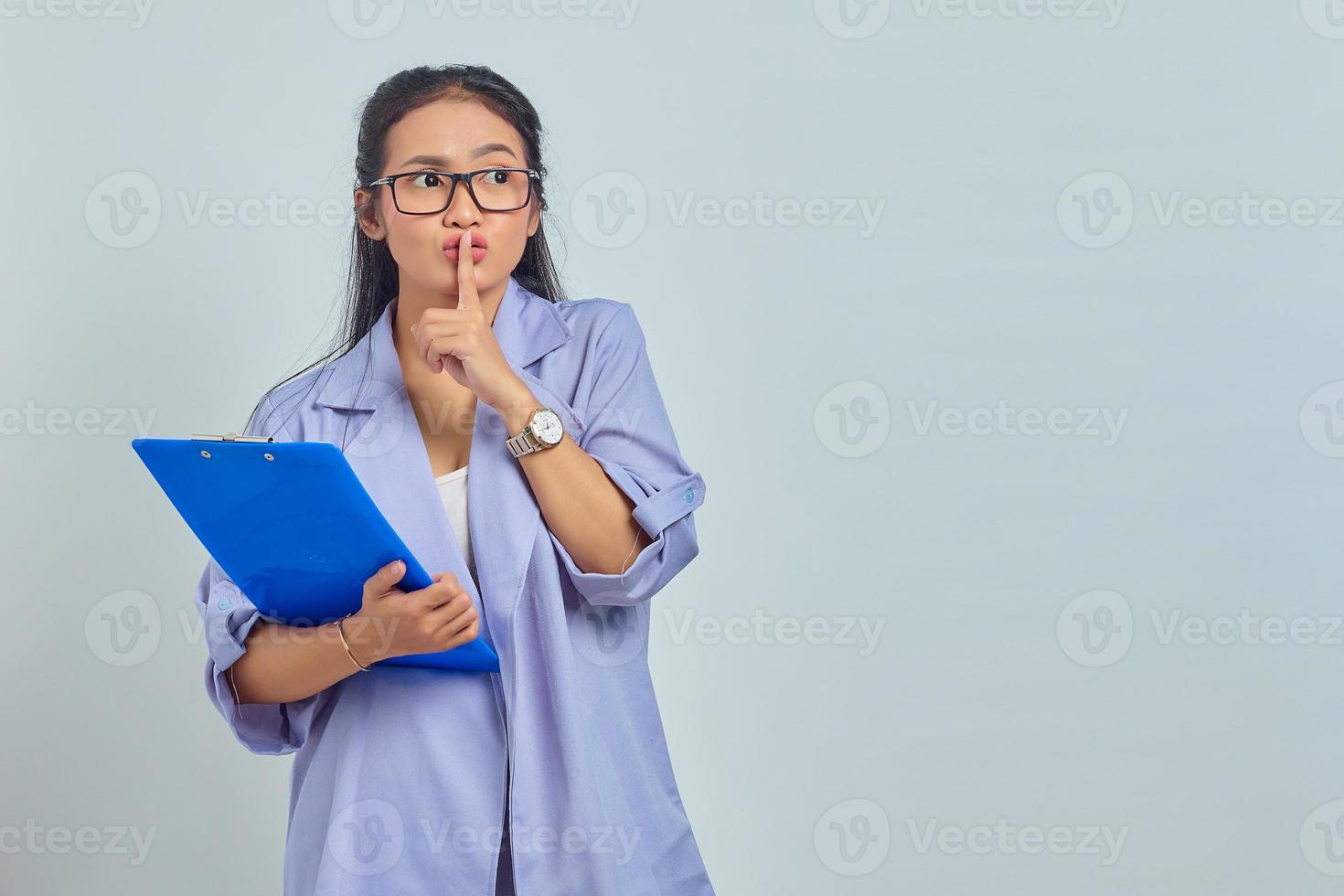 portret van mooie jonge aziatische vrouw die een documentmap vasthoudt en een stil gebaar maakt dat geheim deelt op een paarse achtergrond foto