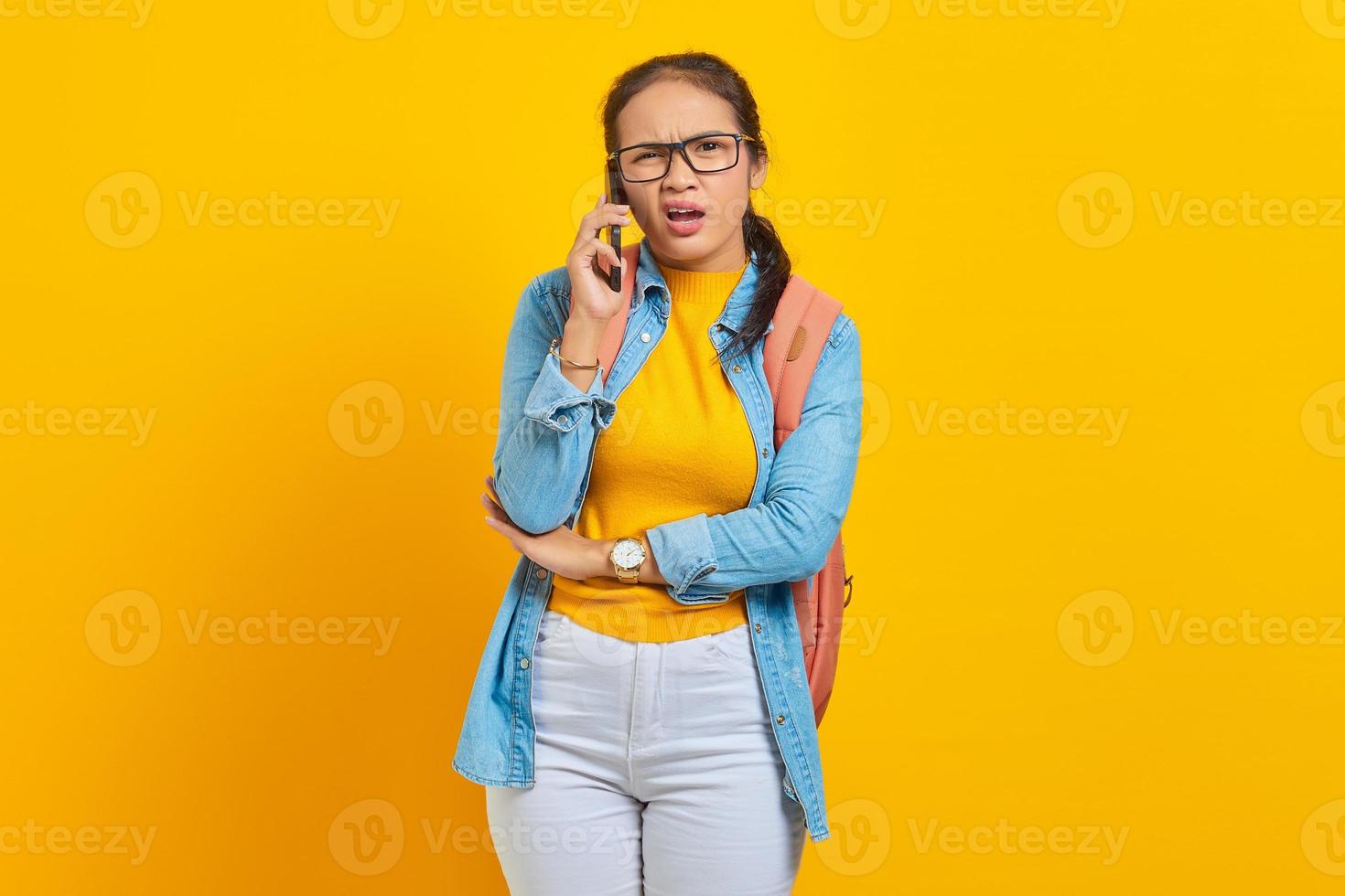 portret van verwarde jonge Aziatische vrouw student in casual kleding met rugzak praten op smartphone en kijken naar camera geïsoleerd op gele achtergrond. onderwijs in universiteitsconcept foto