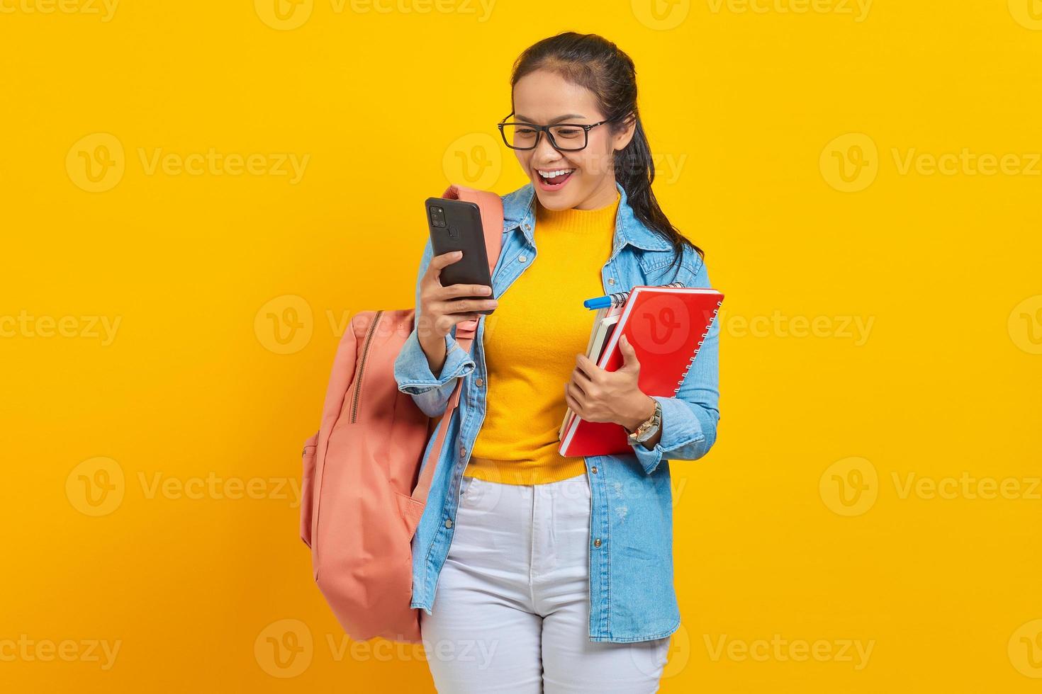 Portret van vrolijke jonge Aziatische vrouw student in denim kleding met rugzak met behulp van mobiele telefoon sms-bericht typen en boeken houden geïsoleerd op gele achtergrond foto