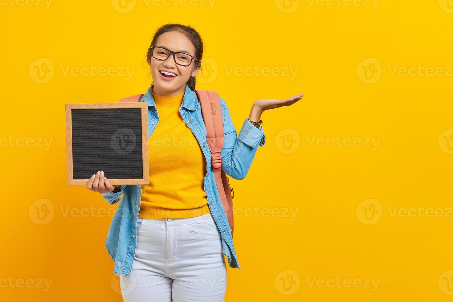 Portret van vrolijke jonge Aziatische vrouw student in casual kleding met rugzak met leeg schoolbord en wijzend op kopie ruimte met palm geïsoleerd op gele achtergrond foto