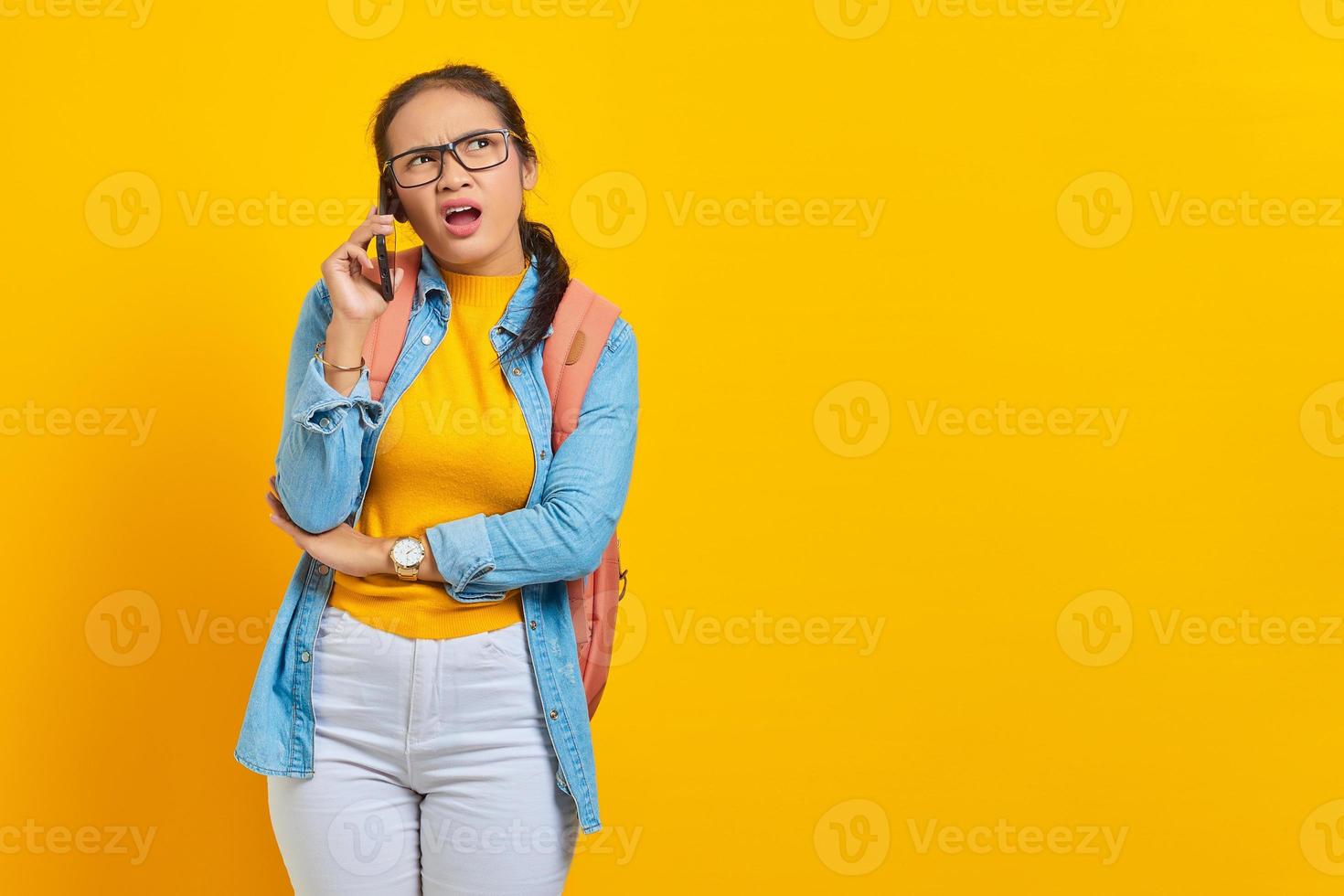 portret van verwarde jonge Aziatische vrouw student in casual kleding met rugzak praten op smartphone en kijken naar kopie ruimte geïsoleerd op gele achtergrond. onderwijs in universiteitsconcept foto