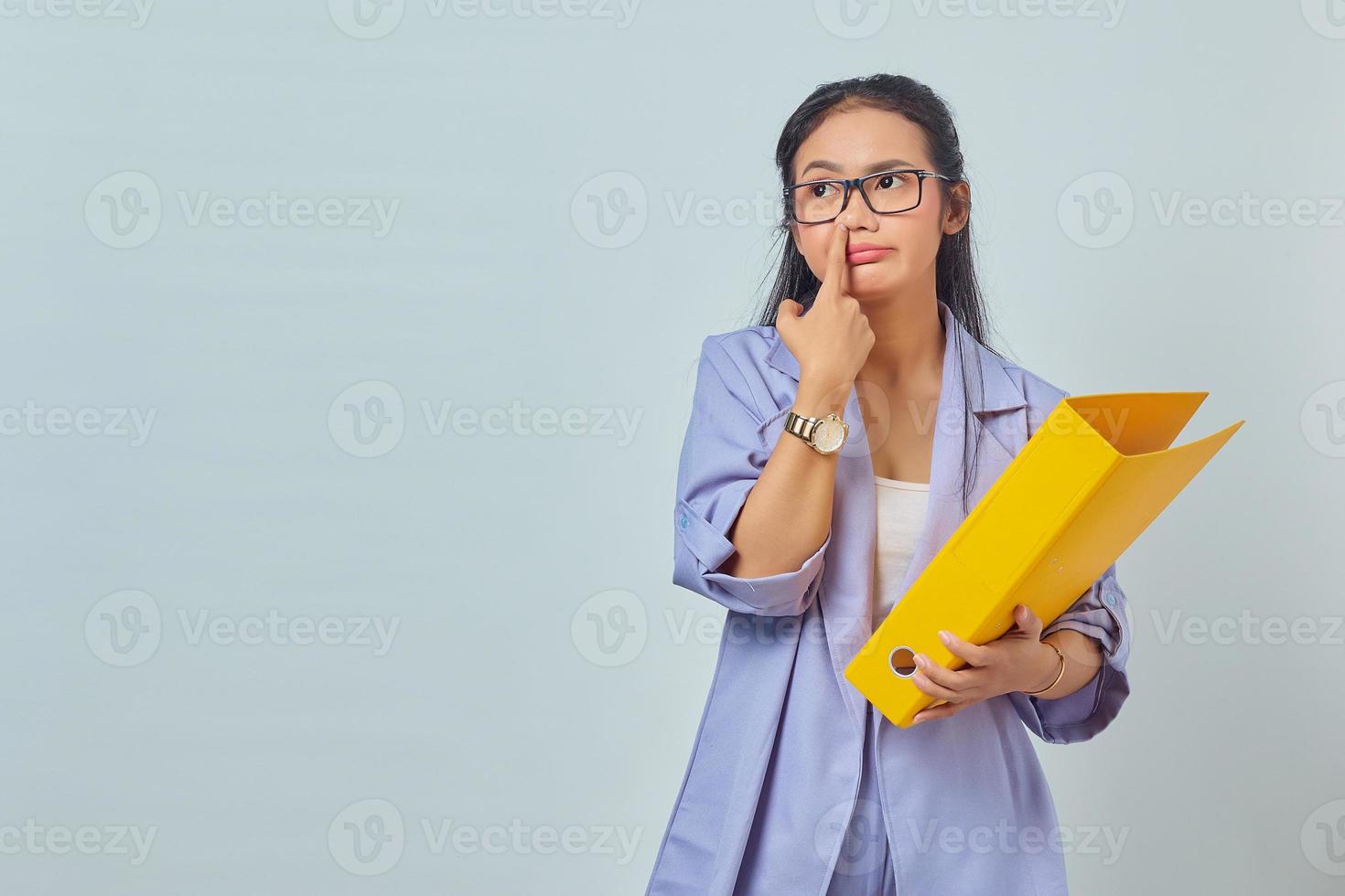 portret van grappige jonge aziatische vrouw die aan iets denkt terwijl ze een gele map draagt en haar neus plukt geïsoleerd op gele achtergrond foto