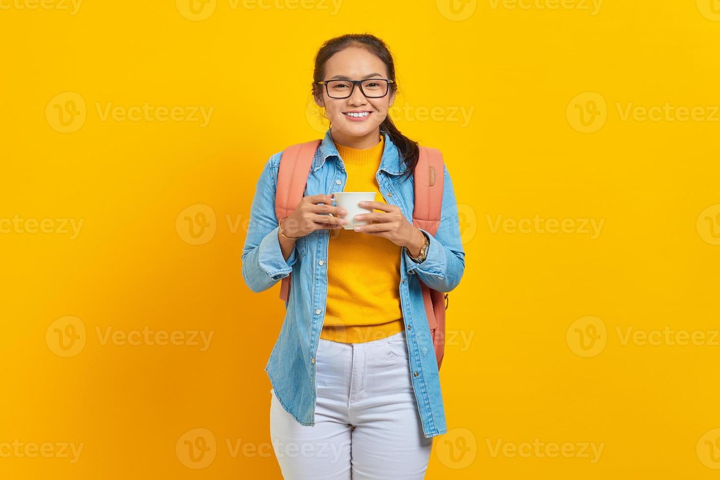 portret van lachende jonge Aziatische vrouw student in denim kleding met rugzak met kopje koffie geïsoleerd op gele achtergrond. onderwijs op de middelbare school universiteit college concept foto