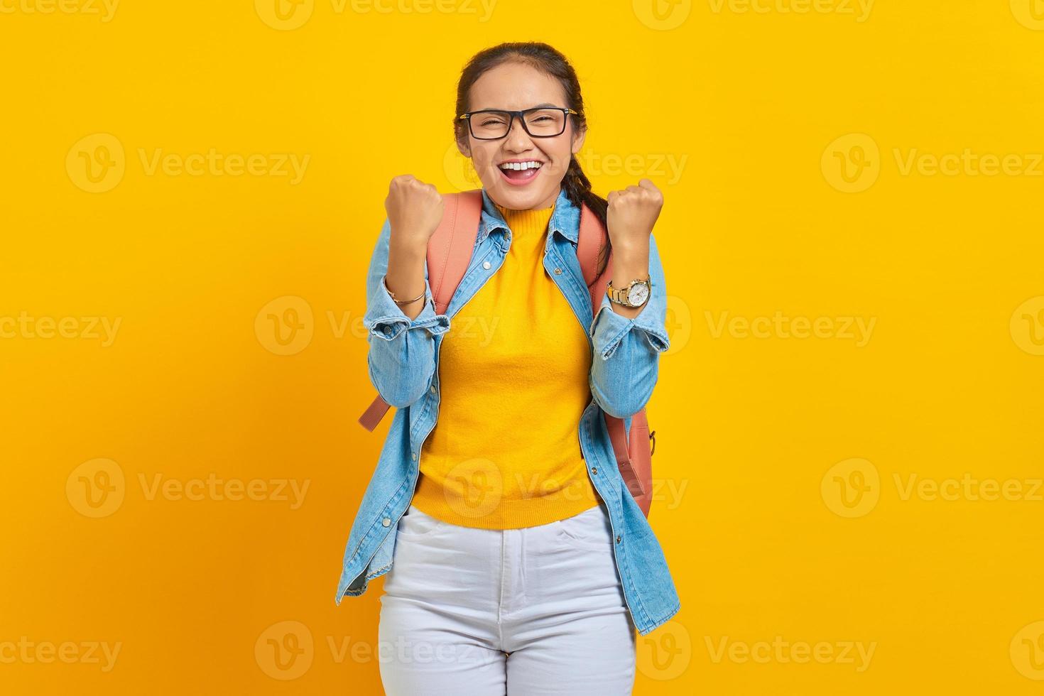 opgewonden jonge Aziatische vrouw student in denim outfit met rugzak staande doen winnende gebaar vieren geïsoleerd op gele achtergrond. onderwijs in universitair concept foto