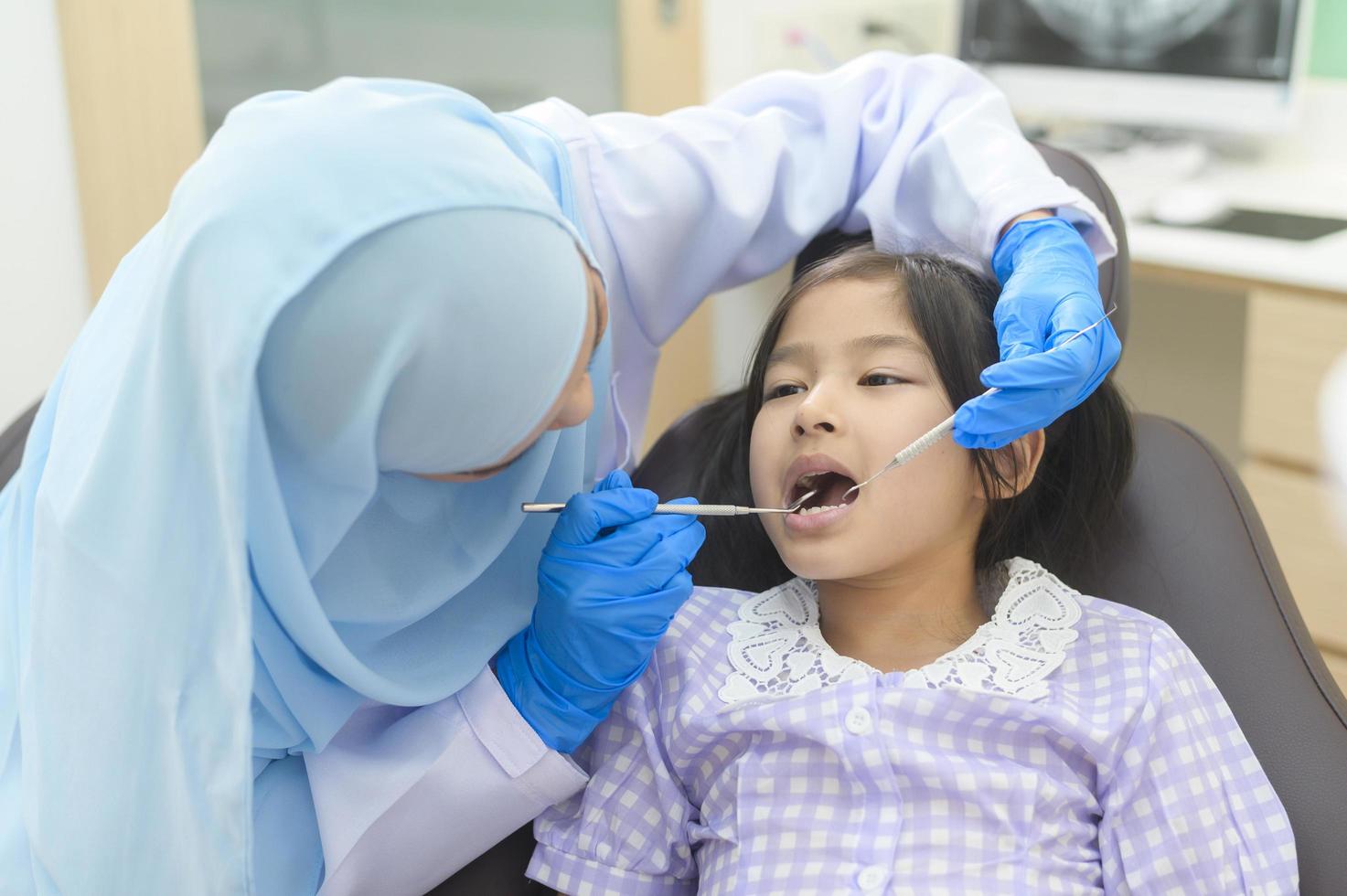 een klein schattig meisje met tanden onderzocht door moslim tandarts in tandheelkundige kliniek, tanden check-up en gezond gebit concept foto