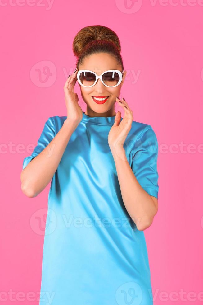 portret van jonge mooie vrouw in blauwe zijden jurk en witte zonnebril op de roze achtergrond foto