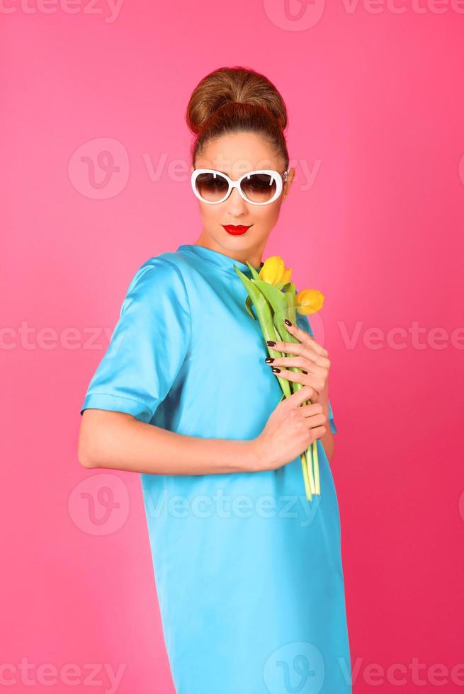 portret van jonge mooie vrouw in blauwe zijden jurk en witte zonnebril met gele tulpen op de roze achtergrond foto