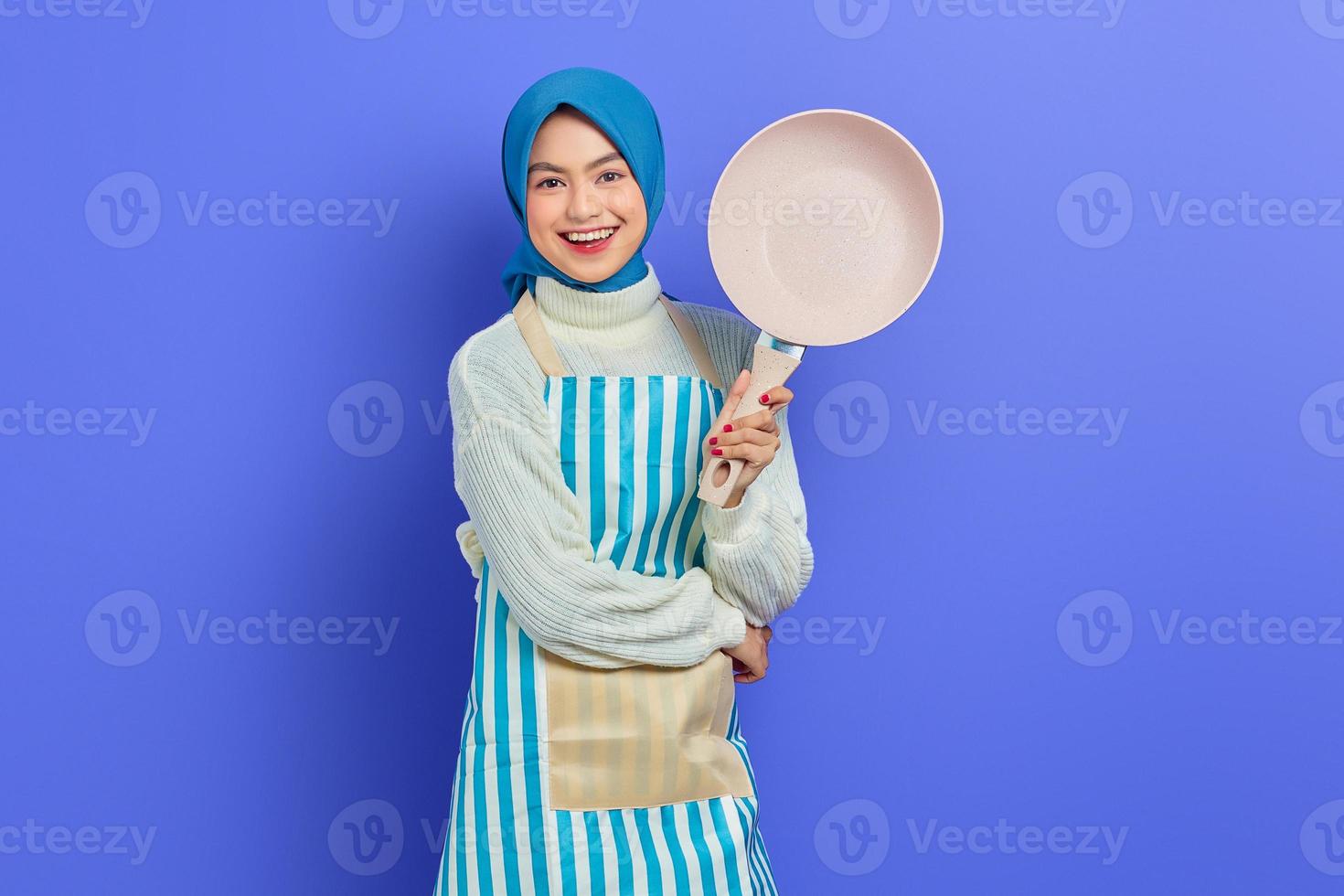 vrolijke mooie aziatische vrouw in schort en hijab houdt pan vast terwijl ze huishoudelijk werk doet geïsoleerd over paarse achtergrond. huishoudconcept foto