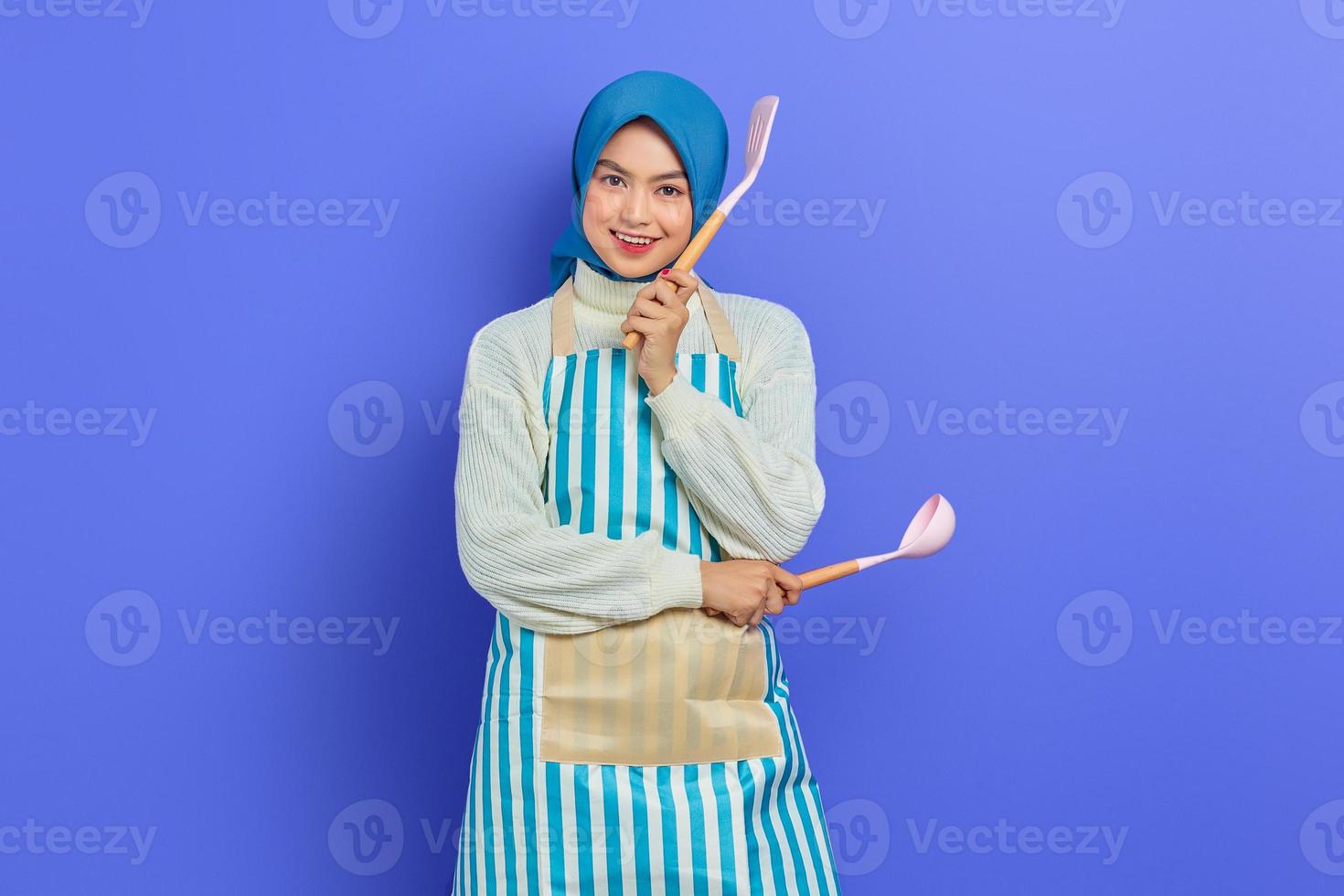 vrolijke jonge Aziatische vrouw huisvrouw in witte schort en trui met lepel soep en spatel terwijl het doen van huishoudelijk werk geïsoleerd op paarse achtergrond studio portret. huishoudconcept foto