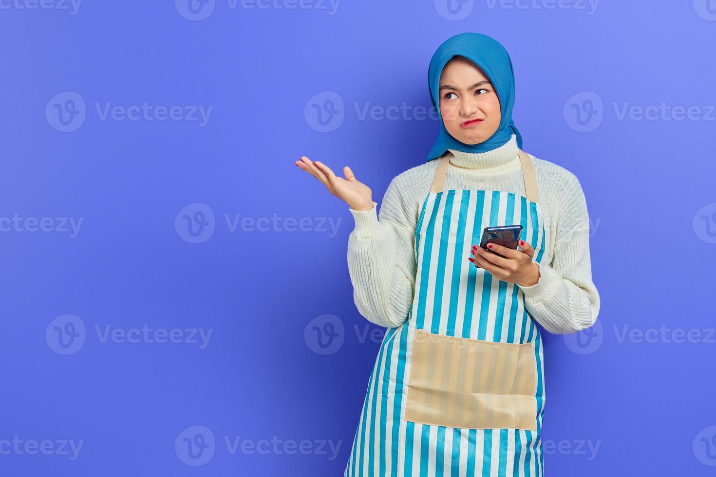 portret van verwarde jonge Aziatische moslimvrouw die hijab en schort draagt die mobiele telefoon houden en exemplaarruimte met handen tonen die over purpere achtergrond worden geïsoleerd. mensen huisvrouw moslim lifestyle concept foto