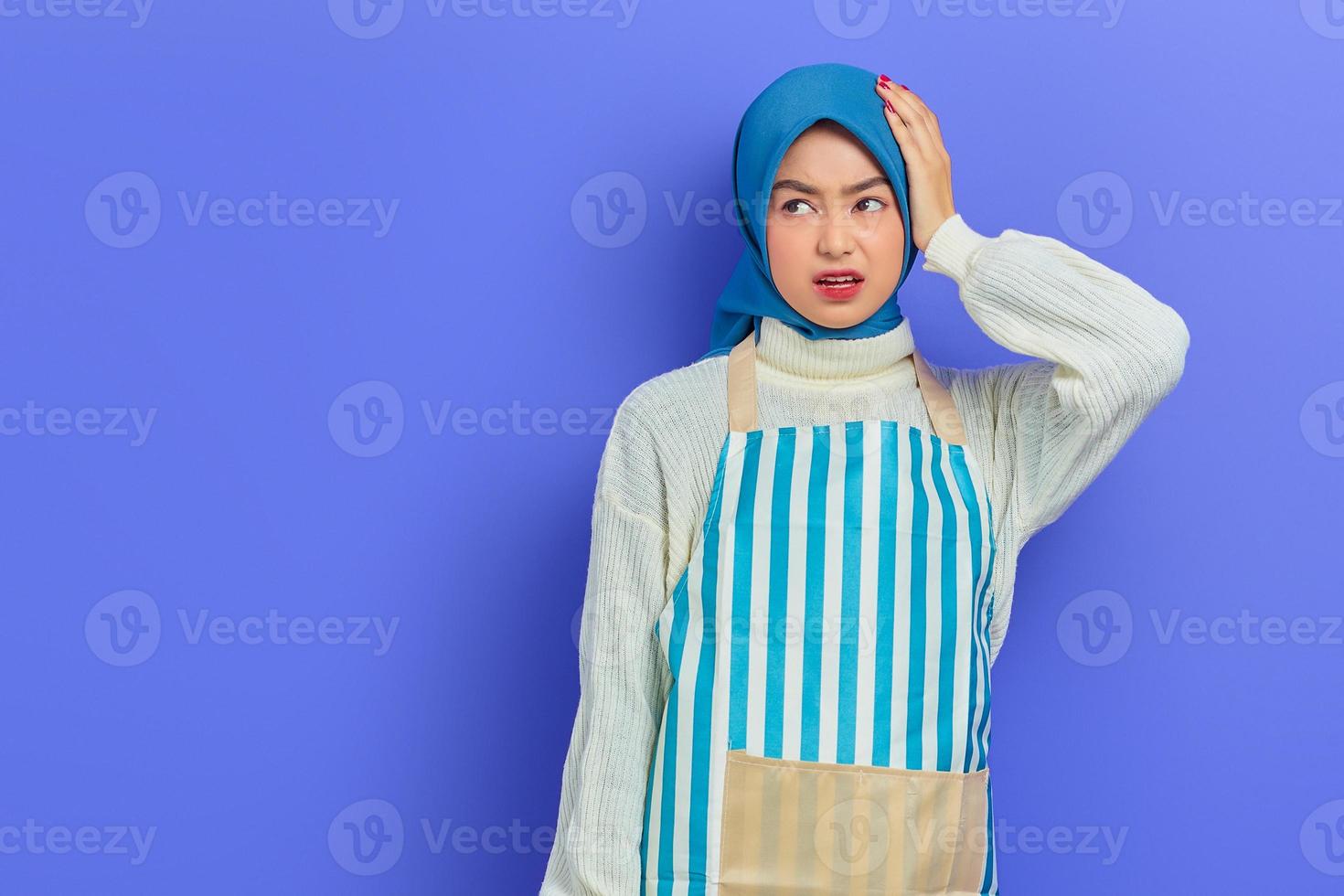 portret van duizelige jonge huisvrouw vrouw in hijab en gestreepte schort opzij kijken terwijl handen op het hoofd geïsoleerd op paarse achtergrond. mensen huisvrouw moslim lifestyle concept foto