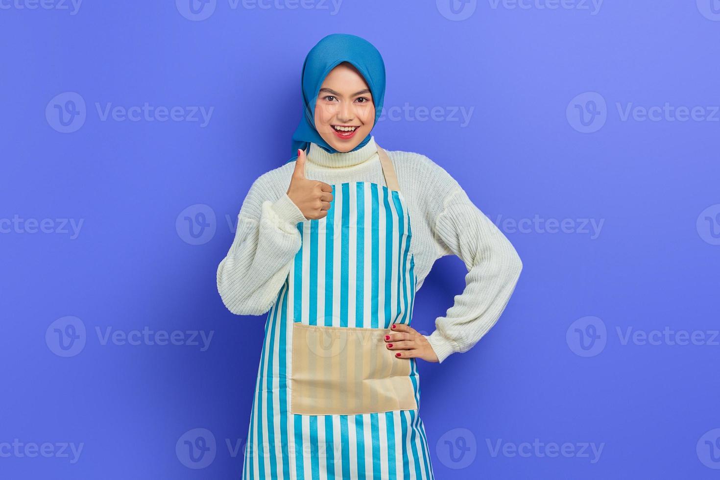 portret van vrolijke jonge huisvrouw vrouw in hijab en gestreepte schort duimen opdagen, op zoek naar camera geïsoleerd op paarse achtergrond. mensen huisvrouw moslim lifestyle concept foto