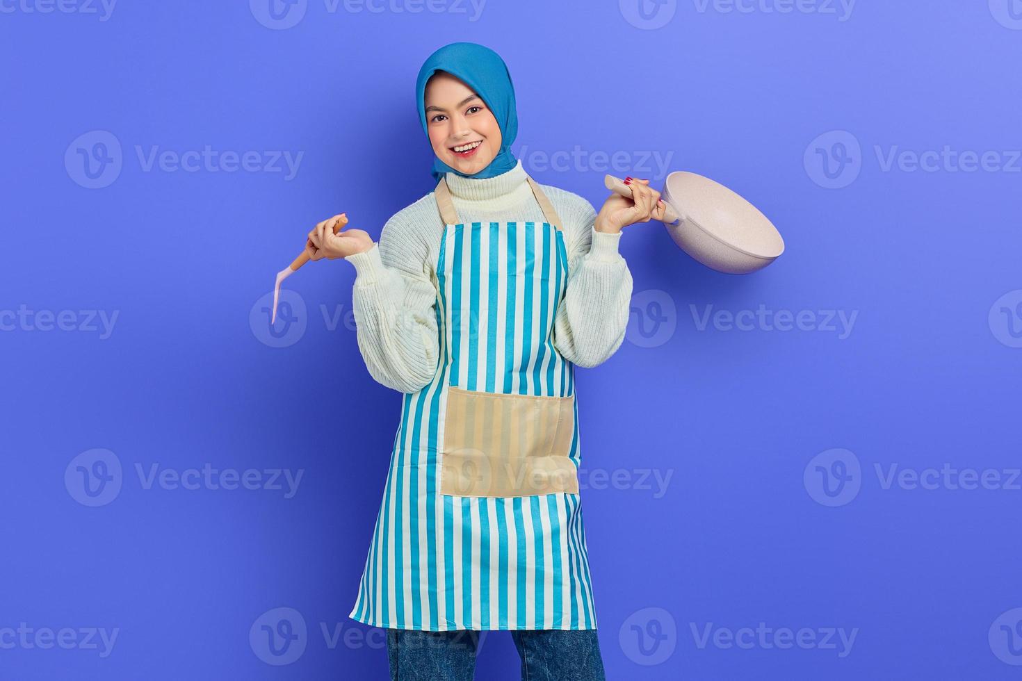 portret van lachende jonge Aziatische moslimvrouw huisvrouw dragen schort met spatel en koekenpan terwijl het doen van huishoudelijk werk geïsoleerd op paarse achtergrond. huishoudconcept foto