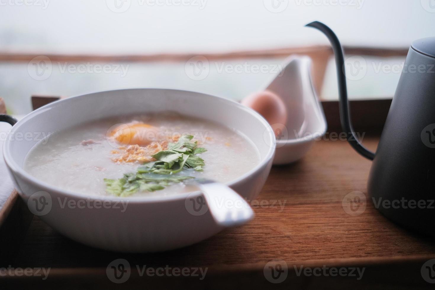 het traditionele Thaise ontbijt in de winter ligt in een houten dienblad op tafel, smakelijke ochtendmaaltijd, warme pap, witte kop gekookte rijst met gebakken knoflook en koriander, ei in een lepel en een waterkoker. foto